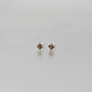 May include: A pair of small stud earrings. Each earring features a round, faceted gemstone with a mix of brown and green hues. The earrings are set on silver posts, and are displayed against a light gray background.