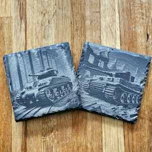 May include: Two black slate coasters featuring engraved images of military tanks. The first coaster depicts a tank in a woodland setting, while the second coaster shows a tank in a more urban environment.