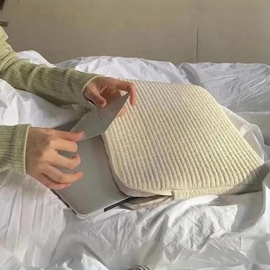 Könnte beinhalten: Eine cremefarbene Laptoptasche mit Steppmuster. Die Tasche ist geöffnet und zeigt einen Laptop. Das Bild wurde in Innenräumen aufgenommen, wobei die Tasche auf einer weißen Oberfläche liegt.