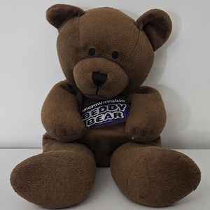 Puede incluir: Un oso de peluche marrón con una nariz negra y un parche azul y morado que dice "Microwavable Beddy Bear". El oso tiene una textura suave y afelpada y está sentado erguido.
