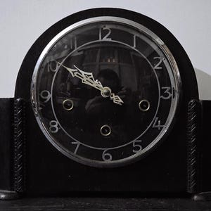 May include: A black Art Deco mantel clock with a circular face and silver trim. The clock face has silver numerals and hands. The clock is rectangular with decorative side panels.