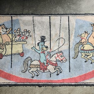 May include: Rectangular rug with a circus theme. Features cartoon animals on carousel horses and in a carriage. The rug has a blue background with a red and beige border, and a fringed edge.