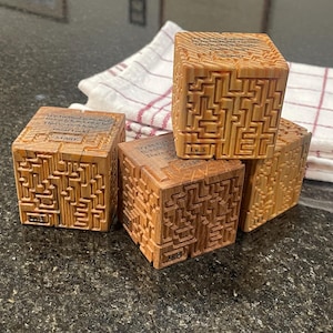 Puede incluir: Cuatro cubos de madera con un diseño laberíntico tallado en sus superficies. Cada cubo tiene texto grabado en un lado. Los cubos están apilados y colocados sobre una encimera oscura, con un paño a rayas blancas y rojas en el fondo.