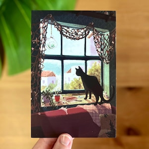 Puede incluir: Postal con una ilustración de un gato negro mirando por una ventana. La ventana tiene una red decorativa y da a una escena costera con edificios blancos y vegetación. Un sofá rojo está en primer plano.
