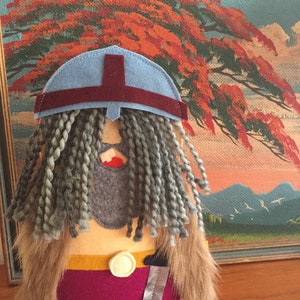 May include: A felt toy of a Viking warrior with a blue helmet, gray braided hair, a burgundy tunic, and a gold belt with a silver knife.