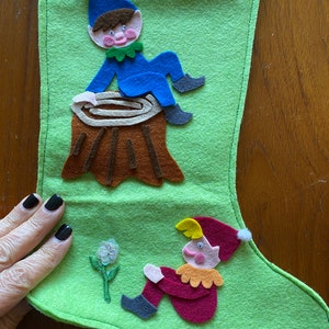 May include: A green felt stocking with two felt elves. One elf is sitting on a brown felt tree stump and the other elf is sitting on the stocking with a white flower in front of them.