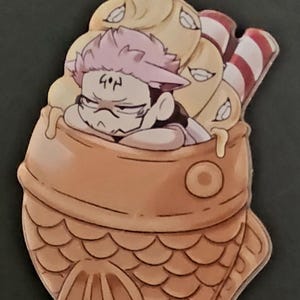 Peut inclure: Un porte-clés doré avec un charm représentant un personnage de dessin animé dans une pâtisserie en forme de poisson. Le charm a un code couleur marron, beige et rose, avec un fermoir doré.
