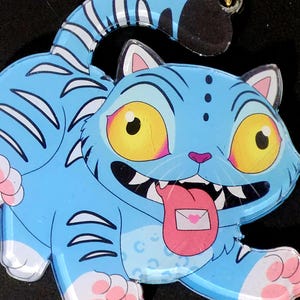 Peut inclure: Un porte-clés en forme de chat bleu avec un fermoir doré. Le chat de dessin animé a des yeux jaunes, des pattes roses et une langue avec une enveloppe rose. Des rayures et des détails blancs ornent le corps du chat.