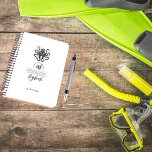 Op de afbeelding: Een wit spiraalgebonden duiklogboek met een octopuslogo en de woorden "DIVING logbook" ligt naast een pen, gele snorkel, masker en vinnen. De duikuitrusting ligt op een houten oppervlak.