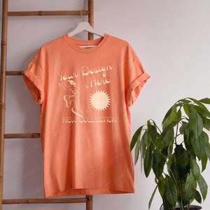 May include: A peach-colored short-sleeve t-shirt featuring a graphic print with the text Your Design Here and NEW COLLECTION, accompanied by a line-art illustration of a person in motion and a stylized sunburst icon on the front center.