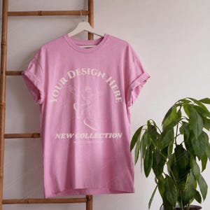May include: A light pink t-shirt with rolled-up sleeves hangs on a white hanger. The shirt features the text "YOUR DESIGN HERE" in a curved arc above a graphic of a person, and "NEW COLLECTION" below. The shirt is displayed against a white wall.