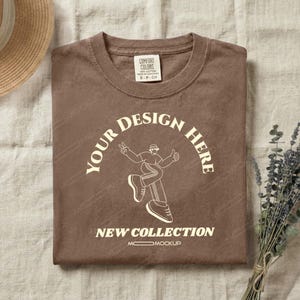 May include: A brown t-shirt with the text "YOUR DESIGN HERE" in an arch, and a graphic of a person. The text "NEW COLLECTION" is below the graphic. The shirt has a tag that says "COMFORT COLORS".