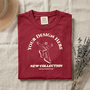 Peut inclure: Un t-shirt bordeaux plié avec un motif graphique blanc représentant une personne avec un signe de paix et un pouce levé. Le texte "YOUR DESIGN HERE" et "NEW COLLECTION" sont également visibles. Un chapeau de paille et une brindille de lavande sont en arrière-plan.