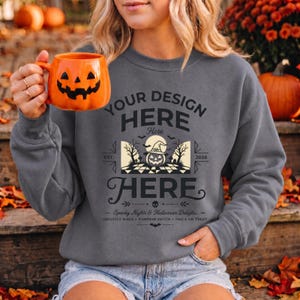 May include: A charcoal gray crewneck sweatshirt featuring a graphic print with the text "YOUR DESIGN HERE" and "HERE". The design includes a central illustration of a jack-o'-lantern wearing a witch hat, trees, and the text "EST. 2026".