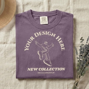Peut inclure: Un t-shirt violet à manches courtes avec un motif crème représentant une personne portant un bob et faisant un signe de paix. Le texte indique YOUR DESIGN HERE et NEW COLLECTION. Le t-shirt possède une étiquette Comfort Colors au col.