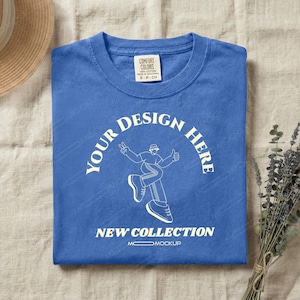 May include: A blue short-sleeve t-shirt with a white graphic print featuring a person in a bucket hat making a peace sign. The text reads YOUR DESIGN HERE and NEW COLLECTION. The shirt has a Comfort Colors label at the inner neckline.