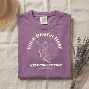 May include: A folded purple t-shirt with the text "YOUR DESIGN HERE" and "NEW COLLECTION" in a white, arched font. A white graphic of a person is in the center. The shirt is on a neutral-colored surface, with a straw hat and lavender sprigs.