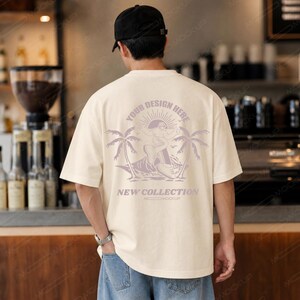 Peut inclure: Un t-shirt à manches courtes blanc cassé avec un motif violet clair au dos. Le design représente une personne faisant du surf entre deux palmiers avec le texte "YOUR DESIGN HERE" et "NEW COLLECTION" imprimé en disposition circulaire.