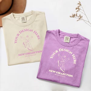 May include: Two crew neck t-shirts, one cream and one purple, featuring the text "YOUR DESIGN HERE" in an arch, a line art illustration of a person dancing, and the text "NEW COLLECTION" with "MOCKUP" below. Both shirts have a neck label.