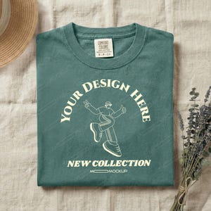 Op de afbeelding: Een opgevouwen tealkleurig T-shirt met een ronde hals. Het shirt heeft de tekst "YOUR DESIGN HERE" in een halve cirkel, met een afbeelding van een persoon. Daaronder staat de tekst "NEW COLLECTION". Een Comfort Colors-label is zichtbaar.