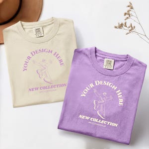 Peut inclure: Deux t-shirts pliés, un crème et un lavande, avec le texte "YOUR DESIGN HERE" et "NEW COLLECTION" dans un design circulaire. Les chemises ont un col rond et une étiquette "COMFORT COLORS".