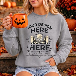Peut inclure: Un sweat-shirt gris à col rond avec une impression graphique noire portant le texte YOUR DESIGN HERE, une illustration centrale d'une citrouille avec un chapeau de sorcière, et le texte Spooky Nights & Halloween Delight, Ghostly Vibes, Pumpkin Patch, Trick or Treat.