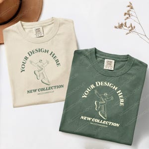 Könnte beinhalten: Zwei T-Shirts mit Rundhalsausschnitt, eines in Beige und eines in Dunkelgrün, jeweils mit einer Grafik einer tanzenden Person und dem Text "YOUR DESIGN HERE" sowie "NEW COLLECTION" auf der Vorderseite. Beide Shirts haben ein Comfort Colors Etikett.