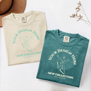 May include: Two folded t-shirts, one cream and one teal, with the text "YOUR DESIGN HERE" and "NEW COLLECTION" printed on them. The shirts are made of a soft material and are perfect for custom designs.