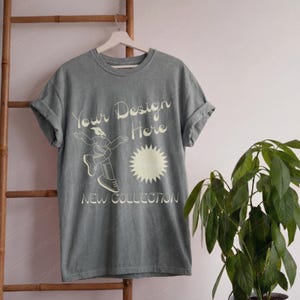 May include: A slate grey short-sleeve t-shirt with a crew neckline and rolled sleeves. The front features cream-coloured text reading Your Design Here and NEW COLLECTION, accompanied by a graphic of a person dancing and a sunburst icon.