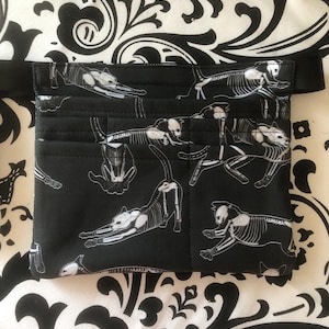 May include: A black waist bag featuring a repeating white graphic print of cat skeletons in various poses. The bag has a rectangular shape with multiple horizontal stitched pockets on the front and an adjustable black strap attached to the sides.