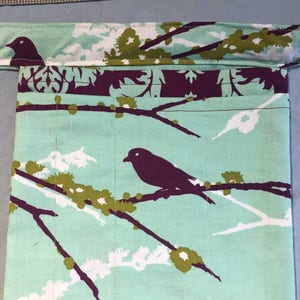 May include: A teal blue fabric pouch with a floral pattern featuring white and green flowers and black birds. The pouch has a purple trim and a brown strap.