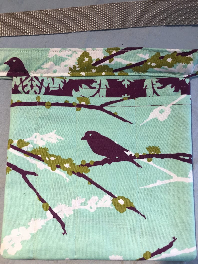 May include: A teal blue fabric pouch with a floral pattern featuring white and green flowers and black birds. The pouch has a purple trim and a brown strap.
