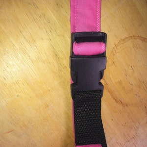 May include: A pink fabric strap with a black plastic buckle. The strap is attached to a black webbing strap.
