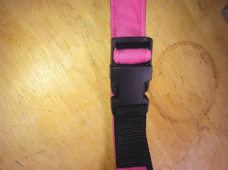 May include: A pink fabric strap with a black plastic buckle. The strap is attached to a black webbing strap.