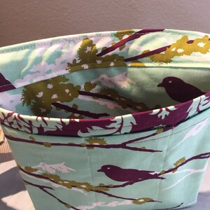 May include: A teal fabric tote bag with a purple and green floral pattern featuring birds. The bag has a purple lining and a strap.