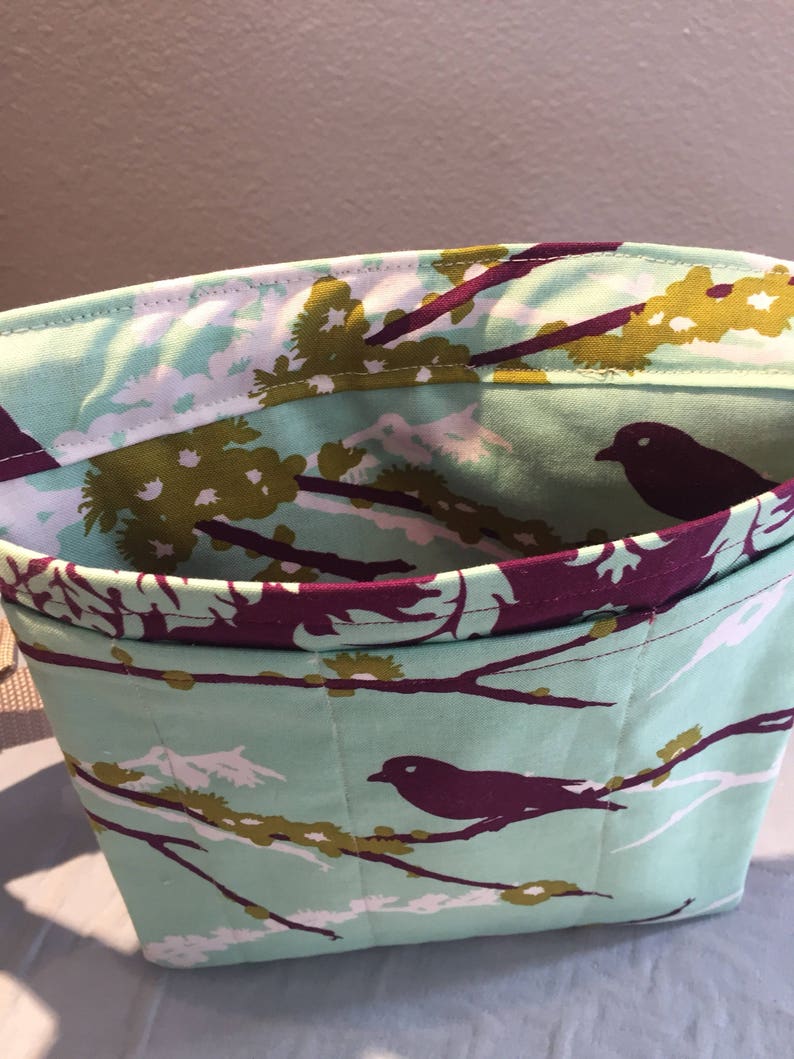 May include: A teal fabric tote bag with a purple and green floral pattern featuring birds. The bag has a purple lining and a strap.