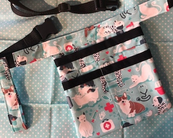 May include: A turquoise blue fabric waist bag with black straps and multiple pockets. The bag has a print of dogs and cats in various poses, including a dog in a bathtub, a cat sleeping, and a dog with a bandage on its leg. The print also includes red crosses, hearts, and bones.