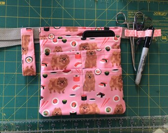 May include: A pink fabric utility belt with multiple pockets, featuring a repeating pattern of brown dogs and sushi rolls. The belt has a black phone pocket and a gray adjustable strap. The belt is lying on a green cutting mat with a ruler and a pair of scissors.