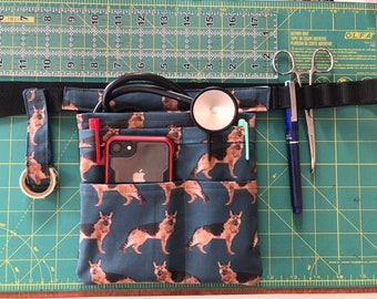 May include: A blue fabric utility belt with multiple pockets, featuring a print of German Shepherd dogs. The belt is equipped with a stethoscope, a cell phone, a pen, and a pair of medical scissors.