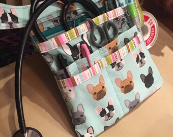 May include: A light blue medical bag with a French bulldog print. The bag has multiple pockets and is filled with medical supplies, including a stethoscope, scissors, and a pen.