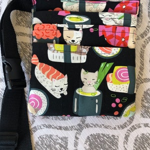 May include: Black fabric pouch with a zipper closure and an adjustable strap. The pouch features a repeating pattern of sushi rolls and cats.