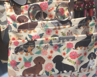 May include: A white canvas bag with a floral and dachshund print. The bag has multiple pockets and is filled with medical supplies, including a stethoscope, scissors, pens, and pencils.