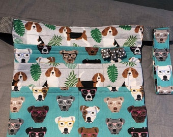 May include: A teal blue fabric pouch with a pattern of dogs wearing glasses. The pouch has multiple pockets and a gray fabric strap with a black buckle. The pouch is made of cotton fabric and has a black trim.