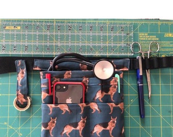May include: A blue fabric utility belt with multiple pockets, featuring a pattern of German Shepherd dogs. The belt is laid out on a green cutting mat with a stethoscope, a pen, and surgical scissors.