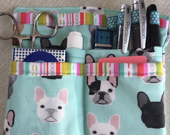 May include: A teal blue fabric organizer with multiple pockets. The organizer has a French bulldog print and is filled with various items, including scissors, pens, a small bottle of hand sanitizer, and a USB drive. The pens are labeled "Precision Inc."