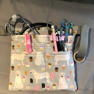 May include: A grey fabric medical utility belt with a white and blue ice cream and dog print. The belt has multiple pockets and is filled with medical supplies, including a stethoscope, scissors, and pencils.