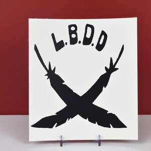 May include: A white square print featuring the text "L.B.D.D" above two crossed black feather-like shapes. The print is displayed on a clear plastic stand against a red background.