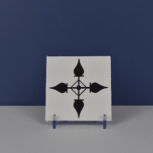 May include: A square, white tile with a black, symmetrical design. The design features four stylized, teardrop-shaped elements pointing outwards from a central square. The tile is displayed on a clear plastic stand against a blue background.