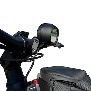 Op de afbeelding: Close-up van een zwart fietsstuur met een gemonteerde koplamp en een kleine zwarte tas. De koplamp is rond en heeft een heldere lens. Het stuur heeft ook een klein beeldscherm en kabels.