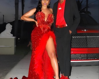 Puede incluir: Una pareja en atuendo formal. La mujer lleva un vestido rojo de lentejuelas con ribete de plumas y una abertura alta. El hombre lleva un traje negro con una camisa roja y zapatos rojos. Están de pie frente a un coche rojo.